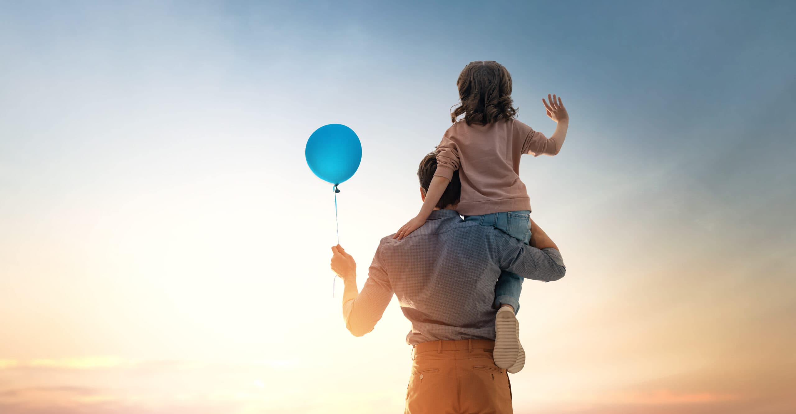 An adule holding a child on their shoulders and holding a blue baloon looking off into the sunset