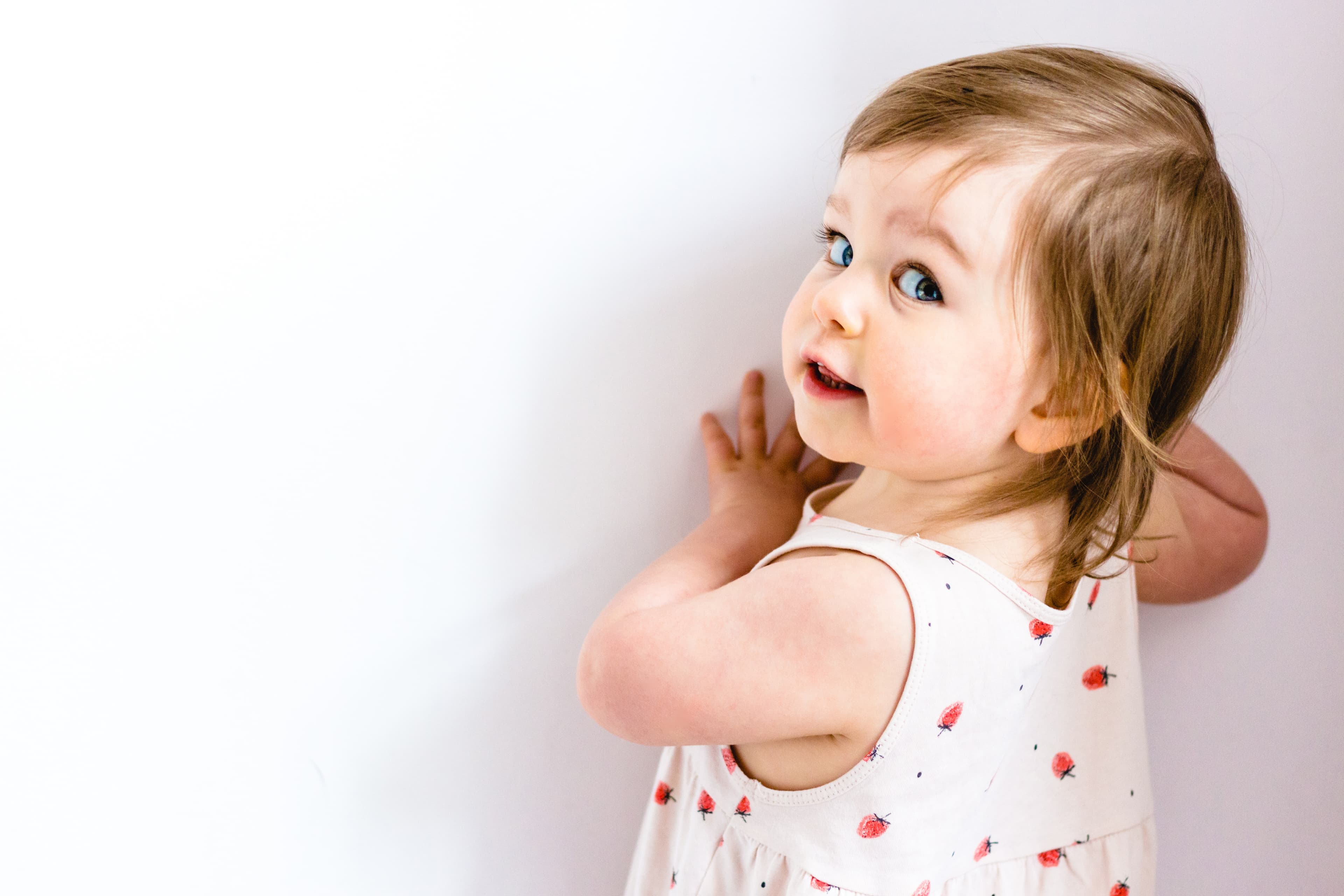 A smiling toddler looks over their shoulder at the camera while holding onto a wall
