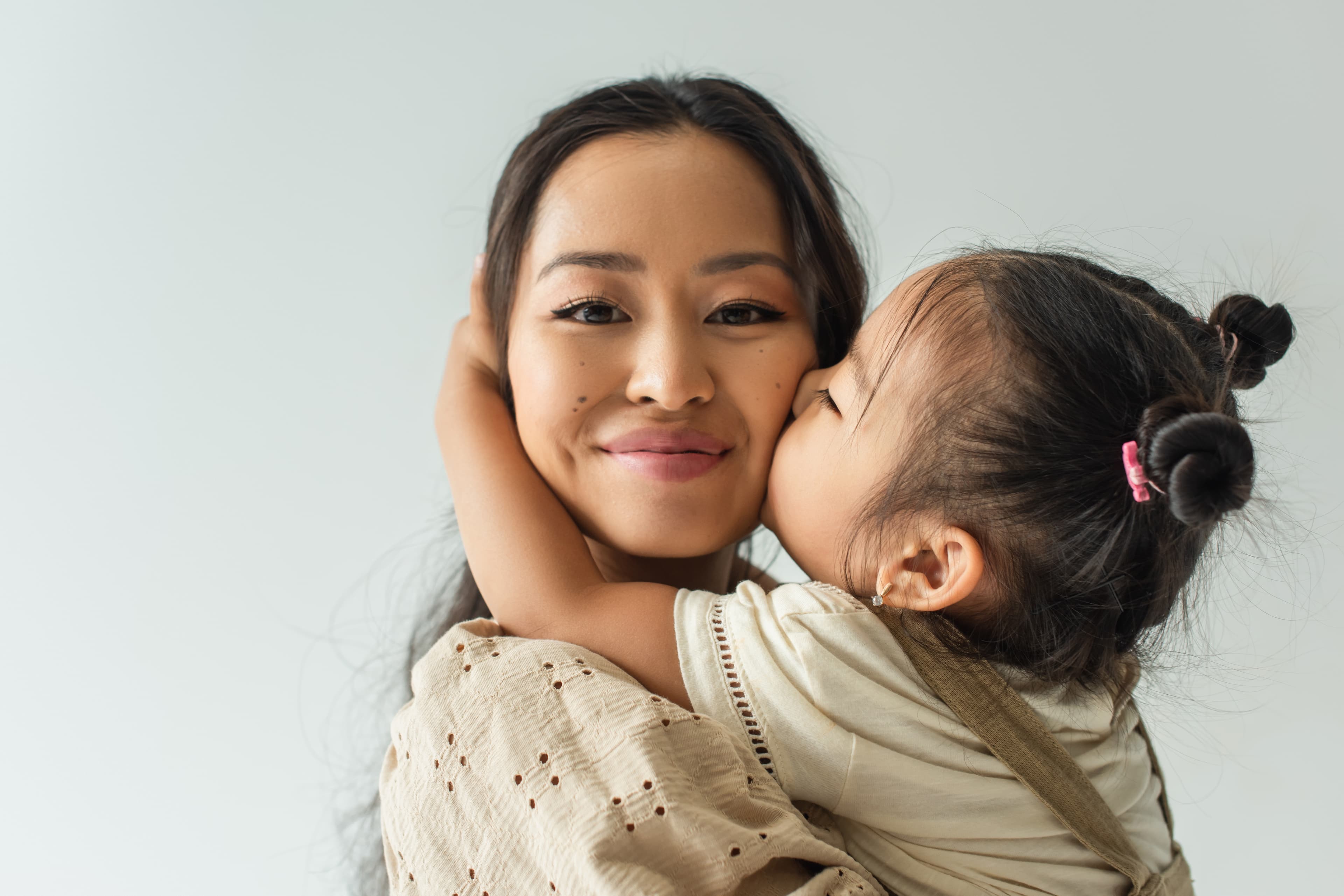 A mother smiles at the camera as her daughter hugs her and kisses her cheek